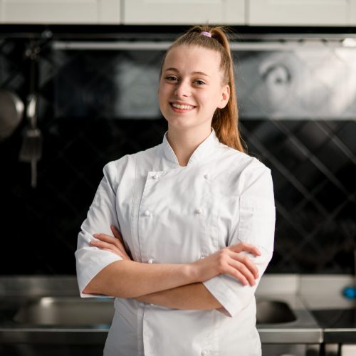 young beautiful smiling woman chef with arms crossed at kitchen young beautiful smiling woman chef with arms crossed wearing in white suit at kitchen. Blurred background
